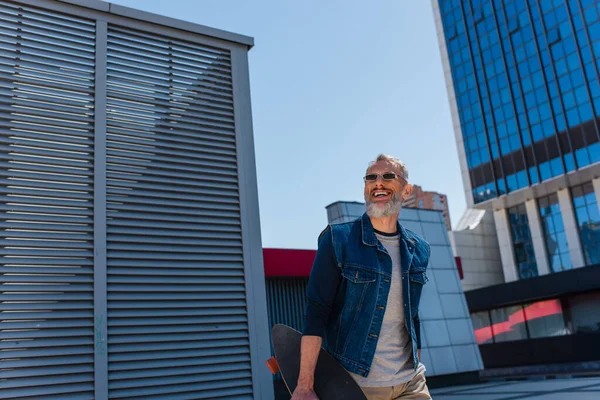 Joyful middle aged man in sunglasses holding longboard on urban street - foto de stock