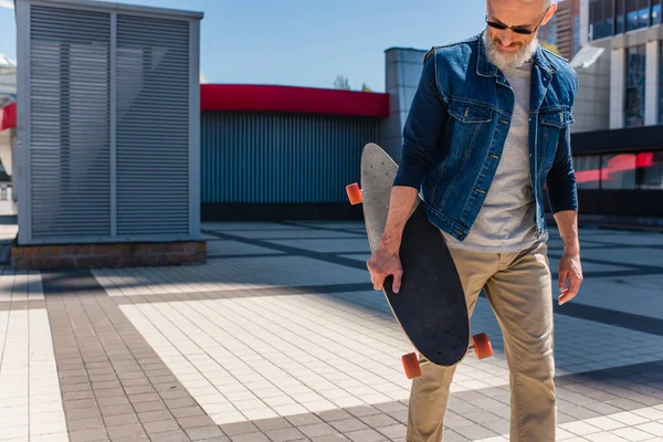 Bearded middle aged man in sunglasses holding longboard on urban street — Stock Photo