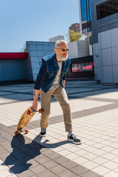 Full length of tattooed middle aged man in sunglasses holding longboard on urban street — Stock Photo