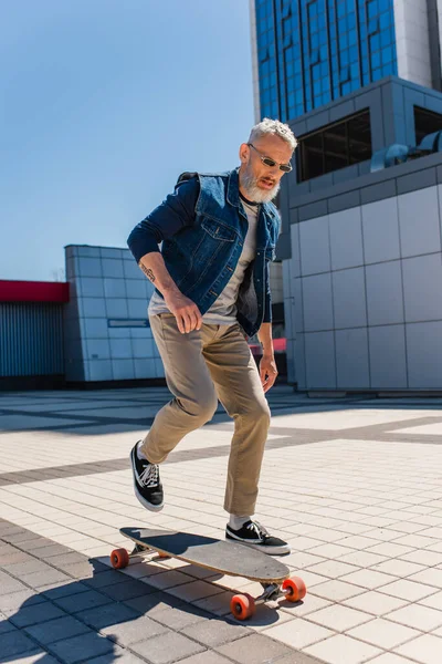 Full length of tattooed middle aged man in sunglasses riding longboard on urban street - foto de stock