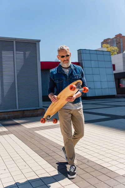 Bearded and mature man in sunglasses holding longboard on urban street — Fotografia de Stock