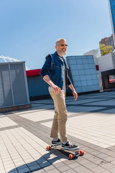 Счастливый и татуированный мужчина средних лет в солнечных очках катается на длинной доске на городской улице — стоковое фото
