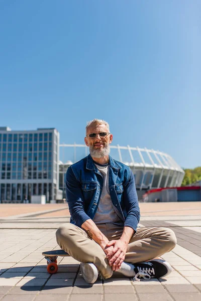 Positive middle aged man in sunglasses sitting on longboard outside — Photo de stock