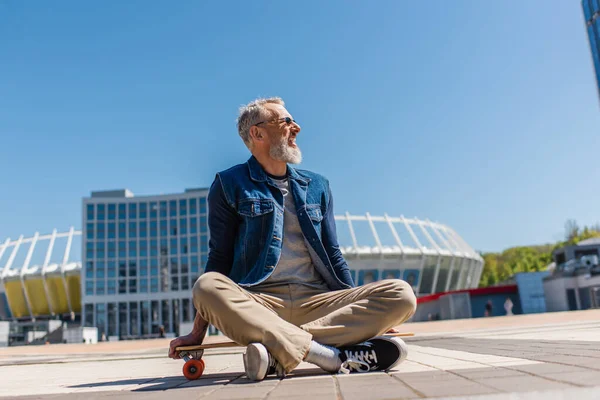 Pleased middle aged man in sunglasses sitting on longboard outside — Stock Photo