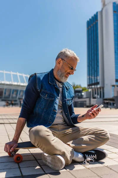 Happy middle aged man in sunglasses sitting on longboard while using cellphone on urban street - foto de stock