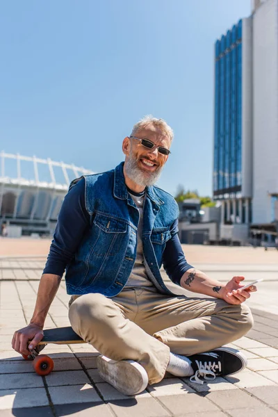 Fröhlicher Mann mittleren Alters mit Sonnenbrille sitzt auf dem Longboard, während er sein Handy auf der Straße benutzt — Stockfoto