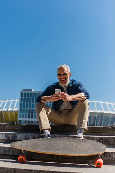 Возбужденный мужчина средних лет в солнцезащитных очках сидит на лестнице рядом с доской, используя мобильный телефон на городской улице — стоковое фото