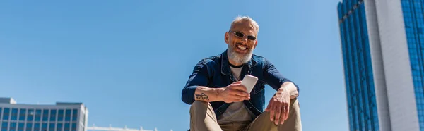 Щасливий чоловік середнього віку в сонцезахисних окулярах сидить і використовує мобільний телефон проти блакитного неба, банер — стокове фото