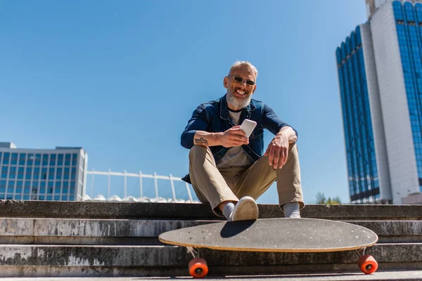 Задоволений чоловік середнього віку в сонцезахисних окулярах, сидячи на сходах біля довгої дошки, використовуючи мобільний телефон на міській вулиці — стокове фото