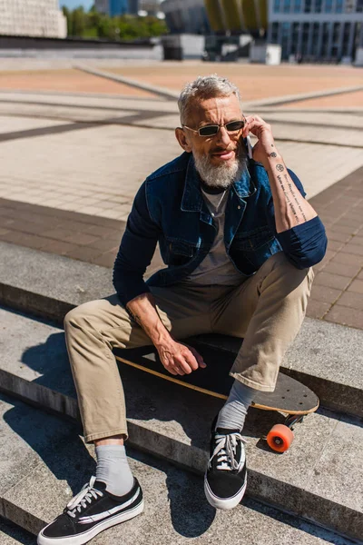 Tattooed middle aged man in sunglasses sitting on stairs near longboard while talking on cellphone on urban street — Stock Photo