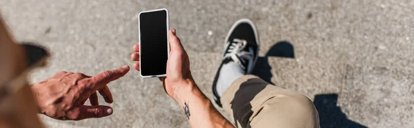 Вид зверху татуйованого зрілого чоловіка, який вказує на смартфон з порожнім екраном, банер — стокове фото