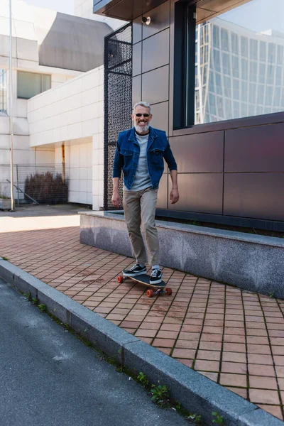 Full length of positive and mature man riding longboard on urban street - foto de stock