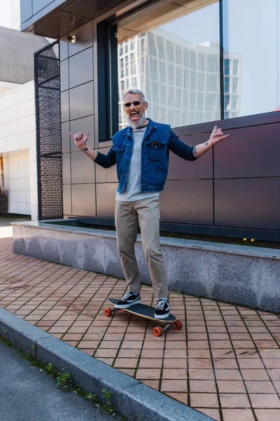 Aufgeregter Mann mit offenem Mund reitet Longboard und zeigt Rockschild auf urbaner Straße — Stockfoto