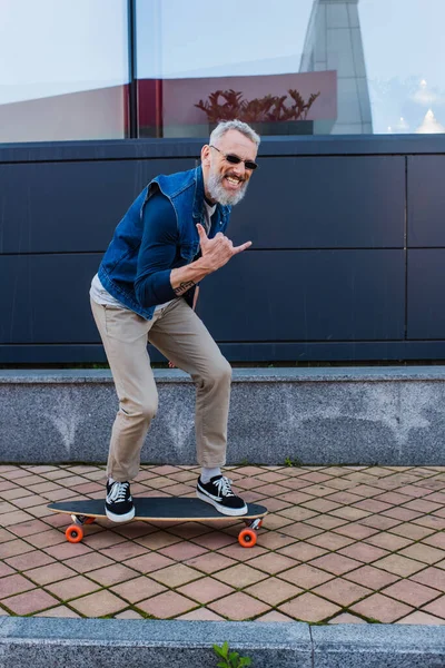 Full length of happy man with open mouth riding longboard and showing rock sign on urban street - foto de stock