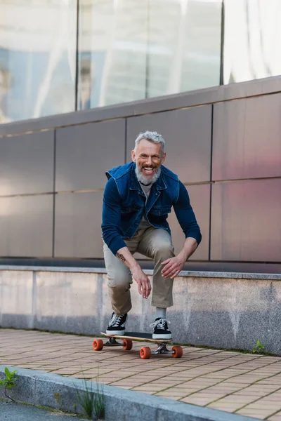 Full length of cheerful and middle aged man riding longboard on urban street — Stock Photo