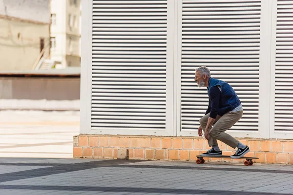 Side view of positive and middle aged man riding longboard on urban street - foto de stock