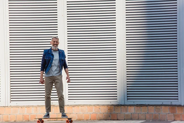 Full length of tattooed and happy middle aged man riding longboard on street — Stock Photo