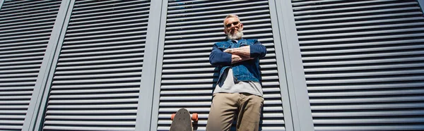 Low angle view of positive and middle aged man standing with crossed arms near longboard on street, banner — Foto stock