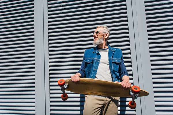 Positive and middle aged man in sunglasses holding longboard — Stock Photo