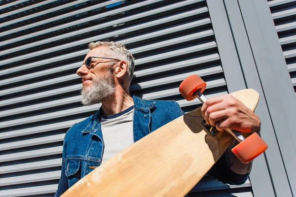 Low angle view of bearded middle aged man in sunglasses holding longboard — Fotografia de Stock