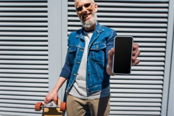 Размытый и довольный мужчина средних лет в солнцезащитных очках, держащий смартфон с чистым экраном и длинной доской — стоковое фото