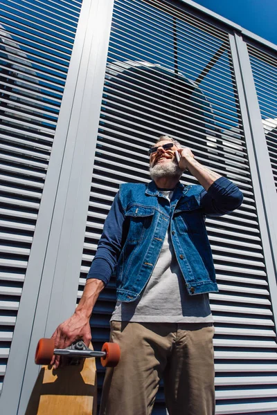 Low angle view of pleased middle aged man in sunglasses talking on smartphone and holding longboard outside — Stock Photo