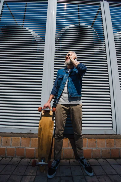 Full length of pleased middle aged man in sunglasses talking on smartphone and holding longboard outside — Stock Photo