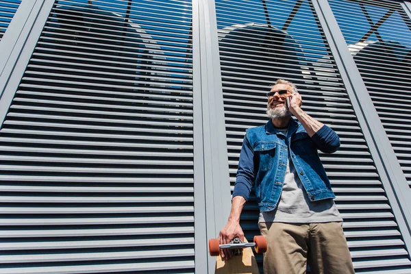 Tiefansicht eines lächelnden Mannes mittleren Alters mit Sonnenbrille, der auf dem Smartphone spricht und Longboard draußen hält — Stockfoto