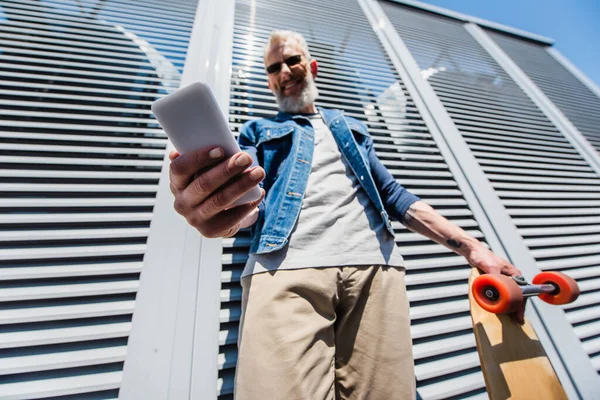 Low angle view of pleased middle aged man in sunglasses using smartphone and holding longboard — Fotografia de Stock