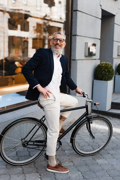 Счастливый бородатый мужчина в очках, позирующий с рукой на бедре и катающийся на велосипеде на городской улице — стоковое фото