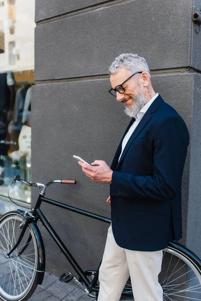 Happy mature man in glasses using smartphone near bicycle on urban street — Photo de stock