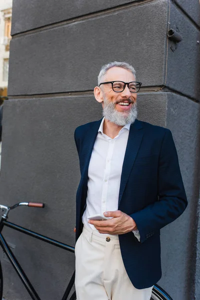 Happy mature man in glasses holding smartphone near bicycle on urban street — Fotografia de Stock