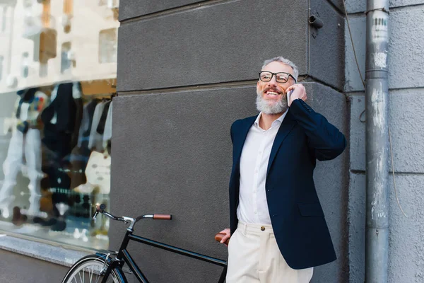 Щасливий чоловік середнього віку в окулярах розмовляє на смартфоні біля велосипеда на міській вулиці — стокове фото