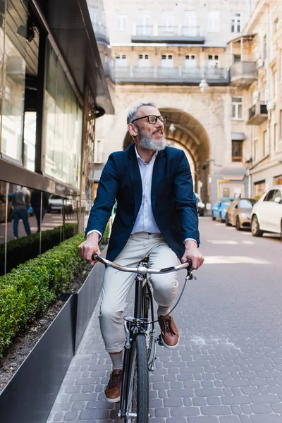 Bearded middle aged man in glasses riding bicycle on modern urban street - foto de stock