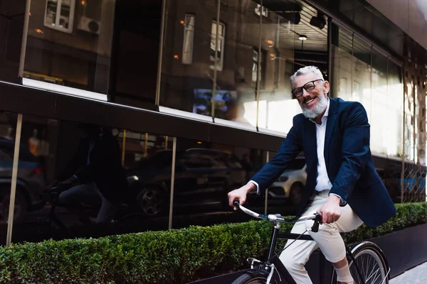 Happy middle aged man in glasses and blazer riding bicycle on modern urban street — Photo de stock