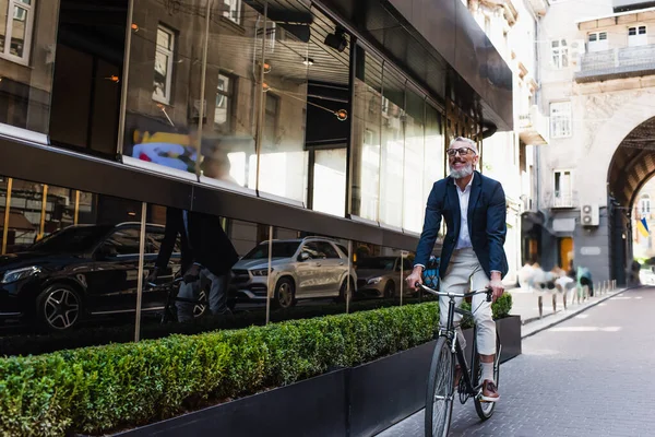 Smiling middle aged man in blazer and glasses riding bicycle on modern urban street — Stock Photo