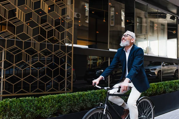 Joyful middle aged man in blazer and glasses riding bicycle on modern urban street - foto de stock