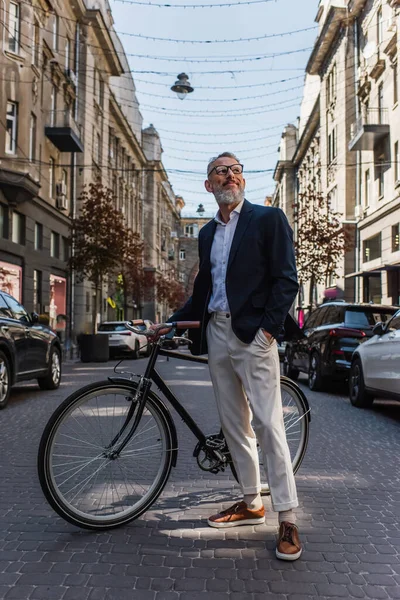 Happy middle aged man in blazer and glasses standing with hand in pocket near bicycle on modern urban street — Fotografia de Stock