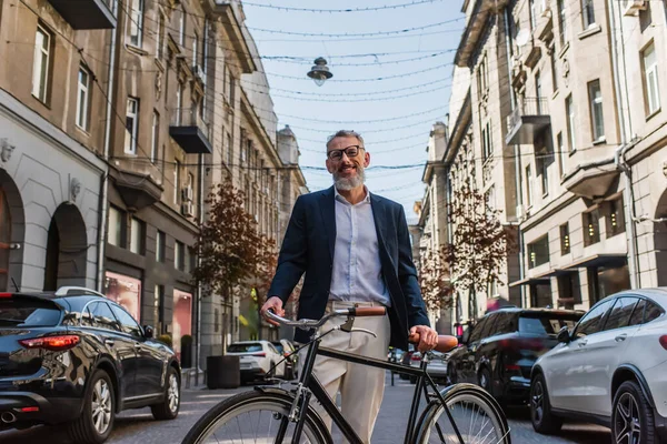 Positive middle aged man in blazer and glasses standing near  bicycle on modern urban street - foto de stock