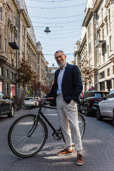 Positive middle aged man in blazer and glasses standing with hand in pocket near bicycle on modern urban street — Photo de stock