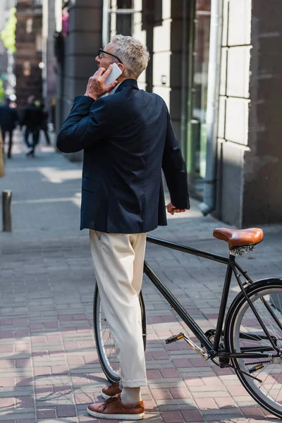 Middle aged man in blazer and glasses talking on smartphone and standing near bicycle on modern urban street — Stock Photo