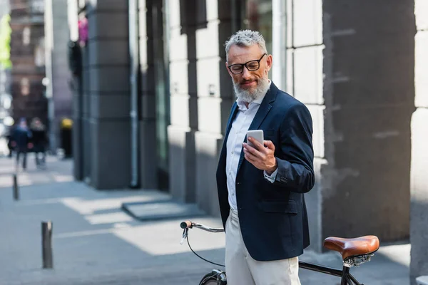 Мужчина среднего возраста в блейзере и очках, смотрящий на смартфон и стоящий возле велосипеда на улице — стоковое фото