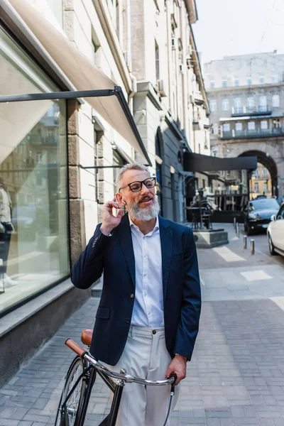 Happy mature man in glasses adjusting earphone near bicycle on street — Fotografia de Stock