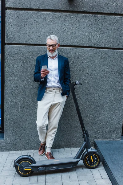 Glücklicher reifer Geschäftsmann im Blazer steht neben E-Scooter und nutzt Smartphone — Stockfoto