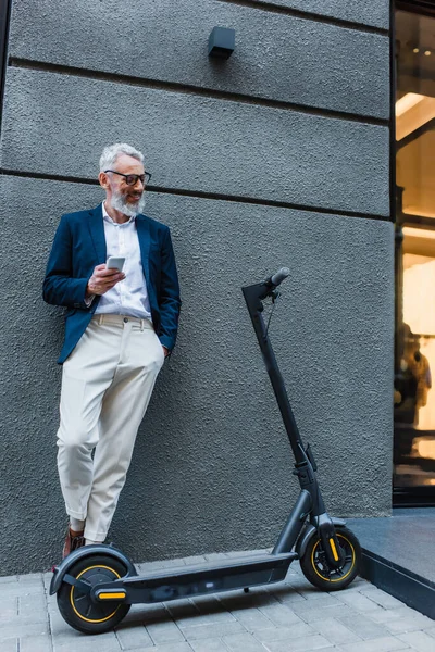 Gut gelaunter Geschäftsmann im Blazer steht neben E-Scooter und nutzt Smartphone — Stockfoto