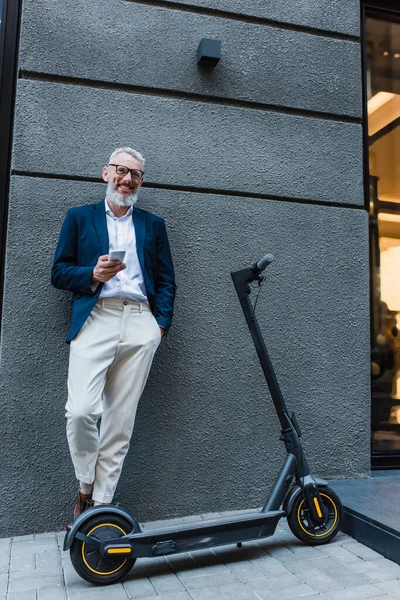 Smiling mature businessman in blazer standing near e-scooter and using smartphone — Stock Photo