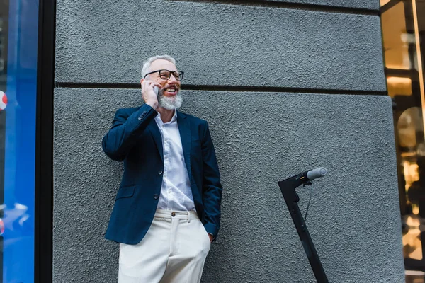 Happy mature businessman in blazer talking on cellphone near e-scooter — Foto stock