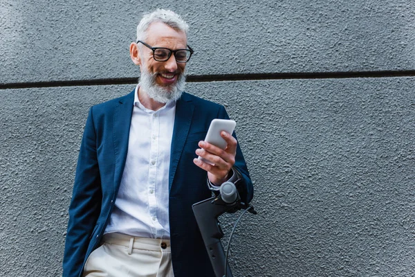 Усміхаючись зрілим бізнесменом у блейзері за допомогою смартфона біля електронного скутера. — стокове фото
