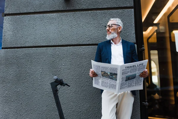 Mature businessman in blazer standing with travel life newspaper near e-scooter — Stock Photo