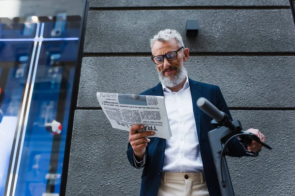 Низький кут зору щасливого зрілого бізнесмена в газеті читання Блейзера поблизу електронного скутера — стокове фото